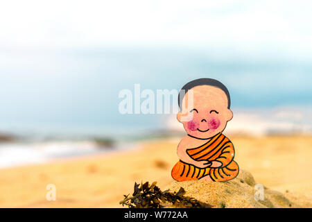 Petit garçon bouddhiste, petit Bouddha, assis sur le sable de la plage . Réjouissez-vous et les sourires dans la position du lotus. Méditation et relaxation Banque D'Images