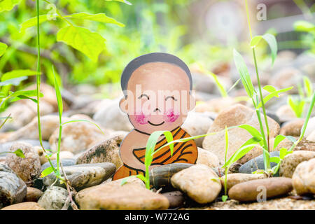 Petit garçon bouddhiste, petit Bouddha, assis sur le sable et pierres sur fond d'herbe verte. et sourires dans la position du lotus. La méditation et Banque D'Images