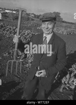 1980s, un homme âgé heureux dans une veste et cravate et casquette plate debout à l'extérieur à son allotissement, Angleterre, Royaume-Uni. Il tient fièrement sa fourchette de jardin, un objet bien utilisé, car trois des dents ou dents métalliques sur la tête de la fourchette sont cassées. Dans sa main gauche sa pipe à tabac. Banque D'Images