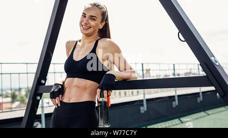 Smiling woman in sportswear leaning on rack puissance tenant une bouteille avec de l'eau Banque D'Images