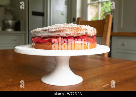 Une éponge victoria maison remplie de fraises fraîches et de crème exposée sur un stand à gâteaux dans un cadre de cuisine confortable. Banque D'Images