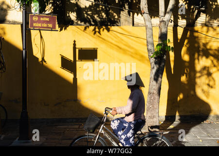 Hoi An, Vietnam - Juin 2019 : Vietnamese woman riding bicycle in vieille ville tomber ombre sur mur jaune Banque D'Images