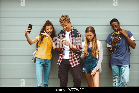 des amis de l'université se tenant dehors regardant à l'aide de leurs téléphones cellulaires. Jeune fille prenant un selfie debout à l'extérieur contre un mur avec ses amis look Banque D'Images