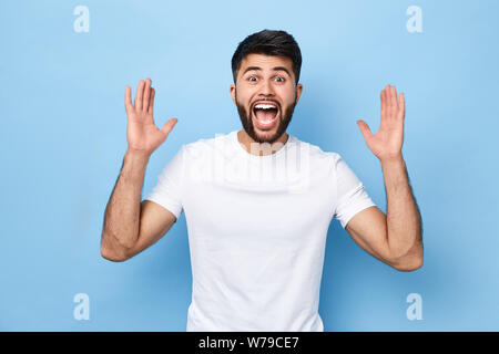 Jeune homme émotionnel dans les tenues de crier avec les bras levés, pour célébrer la victoire, le bonheur et la chance.isolé sur fond bleu. Banque D'Images