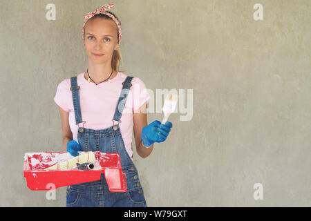 Belle femme avec un pinceau et rouleau pour la peinture. Mur de ciment gris de fond. Banque D'Images