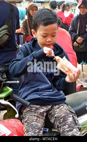 Jakarta, Indonésie - Août 2019 : un garçon mange une saucisse avec stick en face d'un stand d'alimentation de rue. Banque D'Images