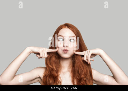 Portrait of happy young redhead woman making crazy face grimaçante et debout contre l'arrière-plan gris Banque D'Images