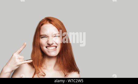 Portrait jeune et mignon adolescente avec de longs cheveux rouges montrant part signer et looking at camera while standing against fond gris. Gesticulant. Carefree Banque D'Images