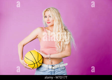 Jeune femme séduisante avec de longs cheveux blonds, beau maquillage, manucure portant haut court rose holding basketball looking at camera with pleasant expressi Banque D'Images