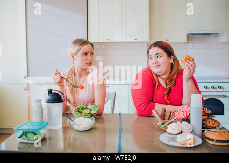 Distrait les jeunes femme bien construite et manger du fromage regarder grande taille mannequin qui tiennent dans la main. burger Happy body positive. Attendus par saines et malsaines Banque D'Images