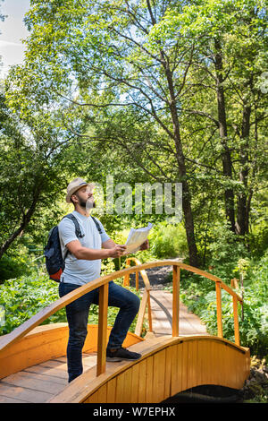 Les jeunes phoques barbus guy millénaire un voyageur dans un chapeau avec un sac à dos se tient sur un pont en bois à la découverte de la région et détermine le chemin sur la carte Banque D'Images