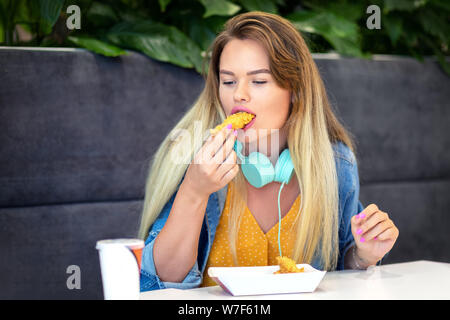 Trendy Young smiling woman eating fast food à bandes croustillantes - Happy hipster girl wearing vêtements et casques holding fried chicken Banque D'Images