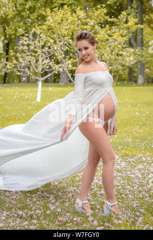 Plein air natural portrait de belle femme enceinte dans l'élaboration dans le vent robe blanche. femme de pétales de fleurs comme la neige Banque D'Images