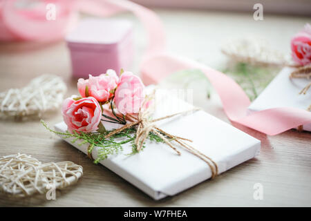 Boîte de cadeau faite à la main enveloppée de papier blanc avec branche de roses. Ruban rose satiné et coeur en bois sur le fond. Banque D'Images