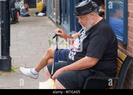 Homme âgé avec une barbe grise assis sur un siège portant un t-shirt punk rock noir et un chapeau en plastique, York, North Yorkshire, Angleterre, Royaume-Uni. Banque D'Images