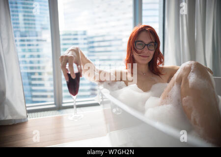 Gingembre une femme couchée dans le bain de mousse et de tenir un verre de vin rouge - vue sur un bâtiment moderne en verre bâtiments de la fenêtres panoramiques Banque D'Images