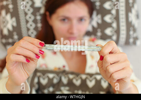 Jeune femme malade au lit de pose, holding classic thermomètre à mercure en face d'elle. Banque D'Images