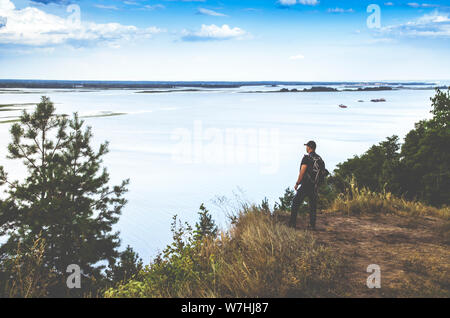 L'homme avec un sac à dos se tient sur le bord d'une falaise surplombant une rivière au milieu d'un ciel bleu Banque D'Images