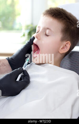 Le dentiste les mains dans des gants en latex noir maintenir l'anesthésie. Le garçon aux yeux clos et la bouche grande ouverte se trouve sur le fauteuil dentaire. Banque D'Images