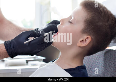 Les mains du dentiste dans les gants en caoutchouc noir tenir une pelle dentaire. L'école un petit garçon est assis sur une chaise avec une bouche ouverte. Le médecin est watchi Banque D'Images