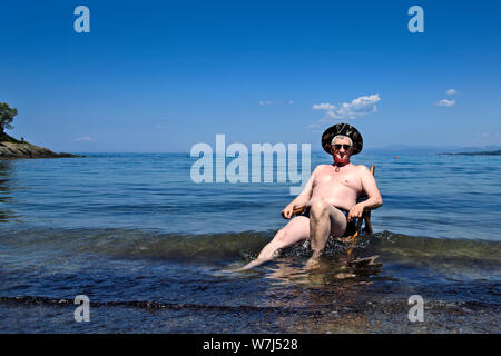 Un homme âgé est assis sur une chaise en bois qui se trouve sur la plage, dans la mer Égée et apprécie le soleil. Banque D'Images