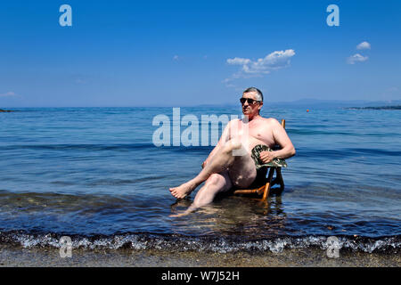 Un homme âgé est assis sur une chaise en bois qui se trouve sur la plage, dans la mer Égée et apprécie le soleil. Banque D'Images