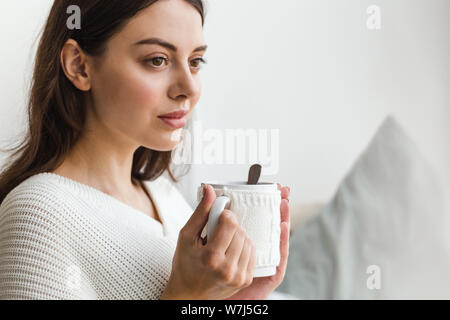 Beau visage d'une jeune fille, une fille en pull blanc est assis sur un canapé avec une tasse de thé chaud dans ses mains Banque D'Images