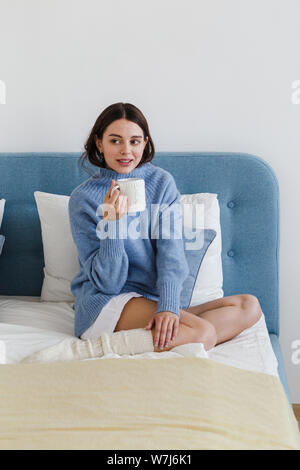 Fille dans un pull bleu dans un style intérieur hygge avec une tasse de thé chaud sa main se repose sur le lit Banque D'Images