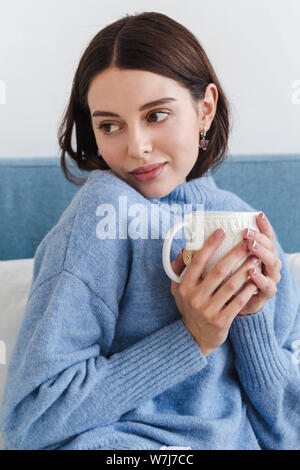 Beau visage d'une jeune fille, une fille en bleu pour homme est assis sur un canapé avec une tasse de thé chaud dans ses mains Banque D'Images