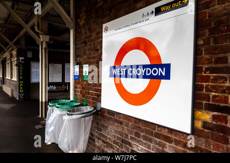 London Overground style railway signent pour West Croydon station. Texte blanc sur une bande bleue, plus de cercle rouge sur fond blanc, positionné contre un mur de briques. Attaché au mur sont des bacs de recyclage pour plastique et papier. Banque D'Images