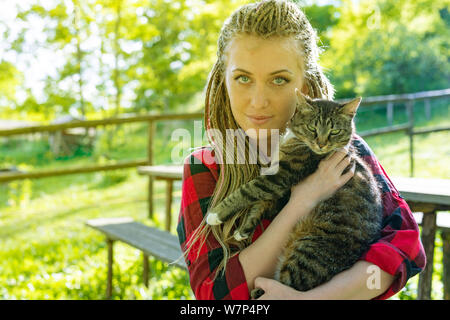 Pretty young woman hugging son chat dans ses bras à l'extérieur dans la douce lumière du soleil d'été à l'extérieur dans le jardin regardant la caméra avec un qu Banque D'Images