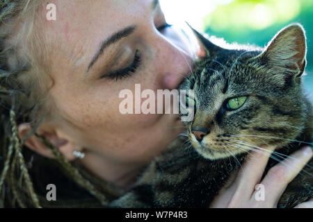 Close up sur happy young woman kissing adorable chat avec les yeux verts. L'arbre vert flou en arrière-plan. Banque D'Images