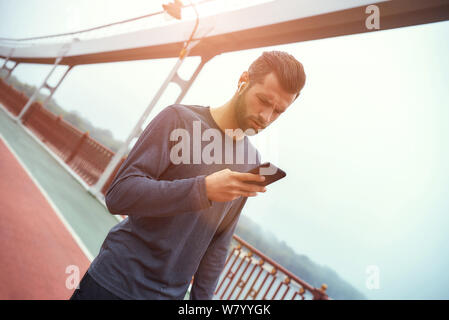 Rester en contact. Jeune et beau barbu homme dans des vêtements de sport et des écouteurs utilisant le téléphone mobile tout en se tenant à l'extérieur sur le pont le matin. Concept numérique. Concept sport. Un mode de vie sain Banque D'Images