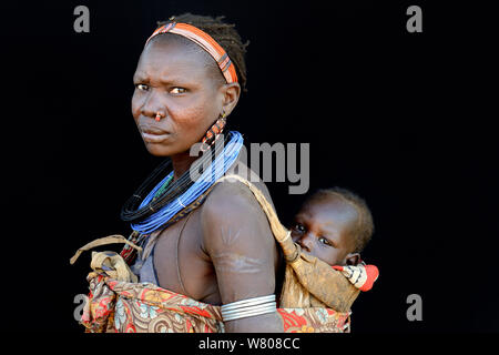 Femme de la tribu Toposas la peau avec scarifications sur le visage et les bras, et portant des bonnets traditionnels et bijoux, portant son bébé sur son dos, vallée de l'Omo, en Ethiopie, en mars 2015. Banque D'Images