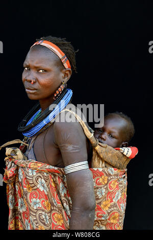 Femme de la tribu Toposas la peau avec scarifications sur le visage et les bras, et portant des bonnets traditionnels et bijoux, portant son bébé sur son dos, vallée de l'Omo, en Ethiopie, en mars 2015. Banque D'Images