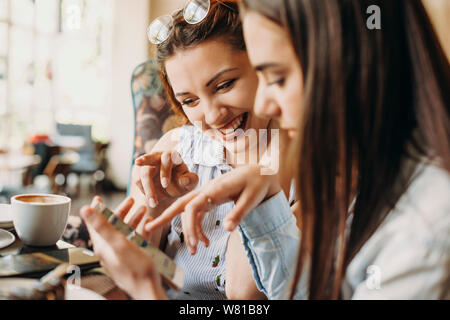 Gros plan d'une belle jeune femme positive du corps aux cheveux rouges montrant quelque chose à ses amies tout en souriant smartphone assis à un bureau. Banque D'Images