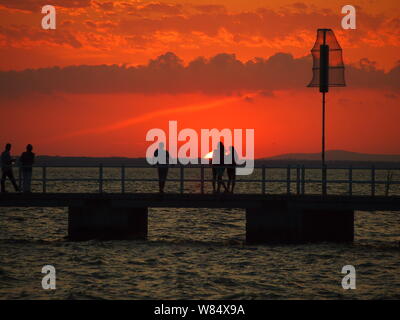Personnes sur un quai silhouetted against orange ciel au coucher du soleil Banque D'Images