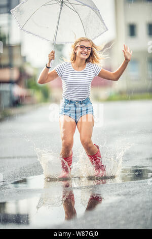 Cheerful girl jumping avec parapluie blanc en rouge en pointillés galoches. Chaude journée d'été après la pluie femme de sauter et de s'éclabousser dans la flaque Banque D'Images