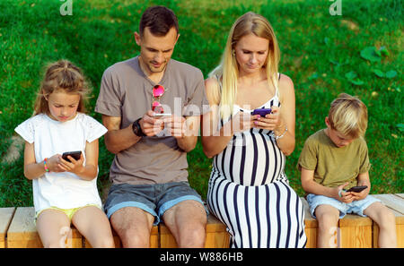 Photo de parents et deux enfants assis sur la clôture en bois avec des téléphones portables dans les mains Banque D'Images