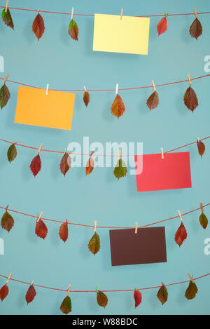 Joyeux automne arrière-plan avec les feuilles d'automne et des feuilles de papier vide de couleur différente, suspendu à des chaînes avec clips en bois, sur un mur bleu. Banque D'Images