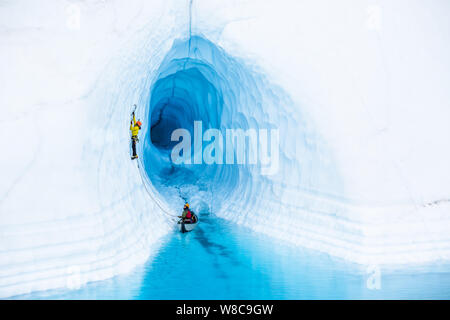 Escalade d'un canot, un grimpeur sur glace mène de l'entrée d'une grotte de glace inondée par une piscine bleu sur le Glacier Matanuska en Alaska. Banque D'Images