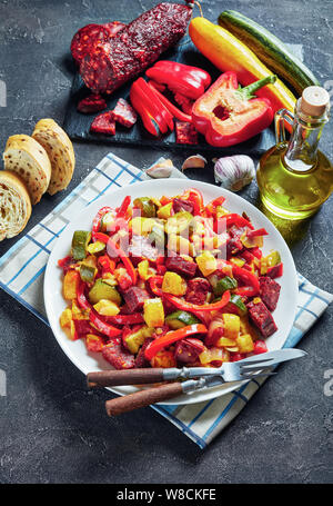 Pisto manchego espagnol - Ragoût aux légumes avec saucisses chorizo frit servi sur une plaque blanche sur une table en béton avec des tranches de pain, vue verticale de Banque D'Images