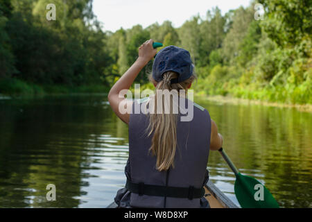 Une jeune fille blonde dans un gilet de l'aviron dans un canoë voile dans les eaux calmes du fleuve par la forêt Banque D'Images