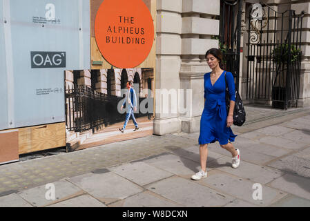 Une dame portant une robe bleu distinctif passe devant un conseil montrant un homme portant une couleur similaire de l'ombre à l'Alpheus Immeuble sur Blomfield Street, un immeuble en cours de rénovation près de la gare de Liverpool Street dans la ville de Londres, le quartier financier de la capitale - aka le Square Mile, le 8 août, à Londres, en Angleterre. L'adresse à 31-35 Blomfield Street est un espace de vente au détail moderne derrière une façade de style édouardien. Banque D'Images