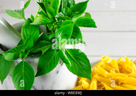 Feuilles de basilic vert dans un mortier en marbre et des pâtes sur une table en bois blanc, close-up Banque D'Images