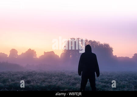 Seul un mystérieux cagoulé, figure debout dans un champ sur un beau matin brumeux au début, en regardant le lever du soleil. Banque D'Images