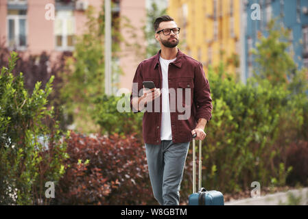 Où je suis beau barbu homme en tenue décontractée et lunettes de vue tirant ses bagages et tenant son smartphone tout en marchant dans la rue de la ville. Concept de voyage. Style de vie Banque D'Images