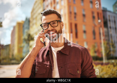 Heureux de vous entendre Portrait d'homme barbu enjoué et beau en tenue décontractée et lunettes de vue parlant au téléphone et souriant en se tenant debout dans la rue. Émotion positive. Numérique. Communication Banque D'Images