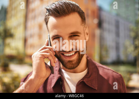 Si bon de vous entendre Portrait d'homme barbu gai et beau parlant par téléphone et souriant en se tenant dans la rue. Bonne nouvelle. Numérique. Communication Banque D'Images