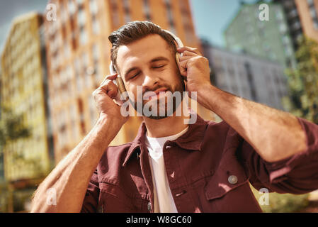 Écouter ma chanson préférée. Beau jeune homme barbu dans un casque d'écoute, écoutant de la musique et gardant les yeux fermés tout en étant debout dans la rue. Inspiration. Concept de musique. Son Banque D'Images